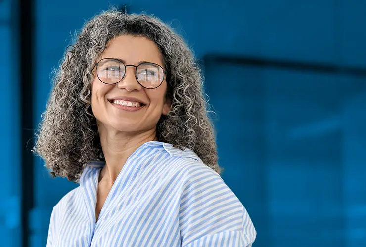 Smiling woman with curly gray hair, glasses, and a striped shirt, looking right against a blurred blue background.
