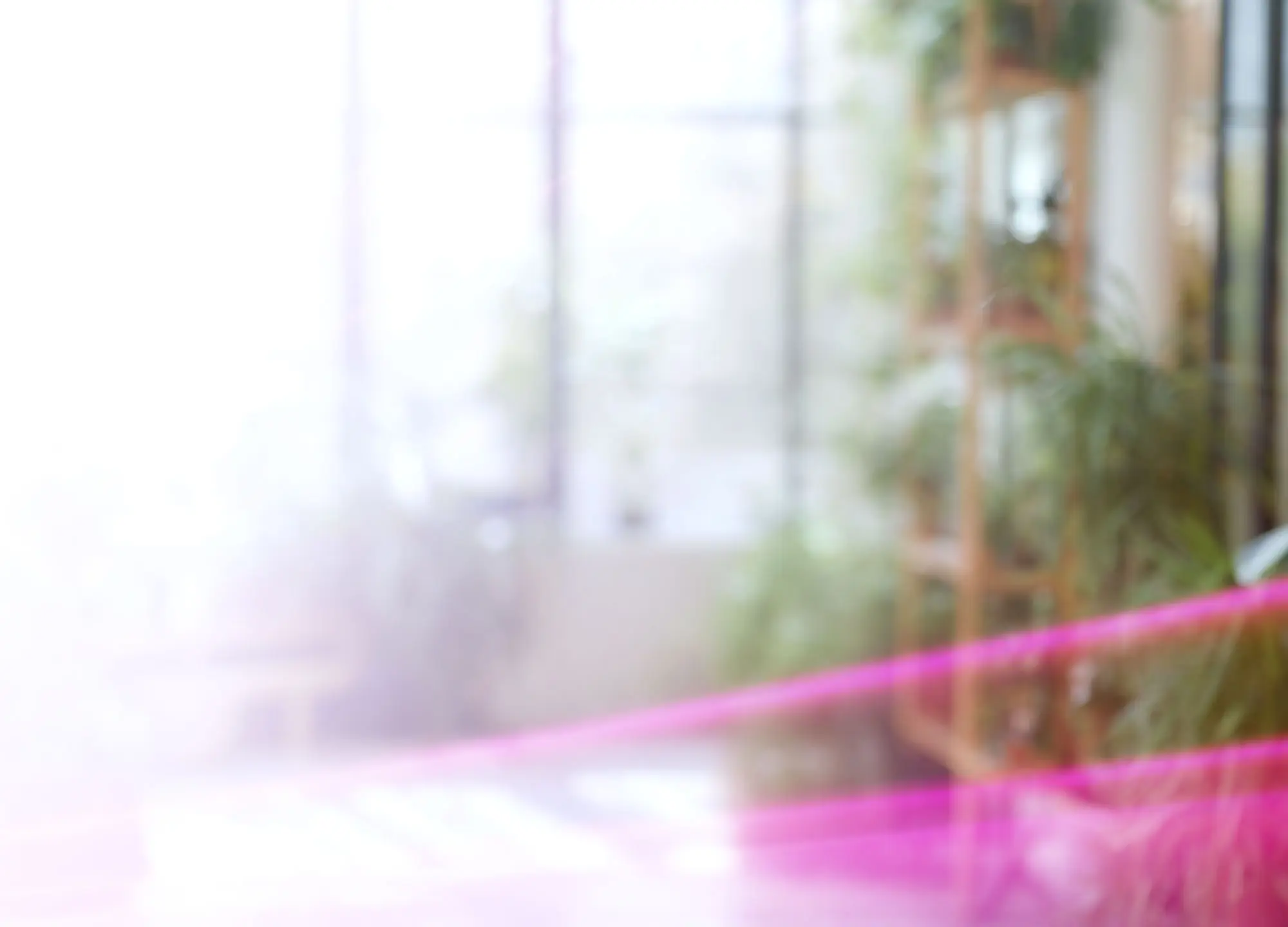 Blurred indoor room with bright background light, faint green plants, and a prominent pink ribbon.
