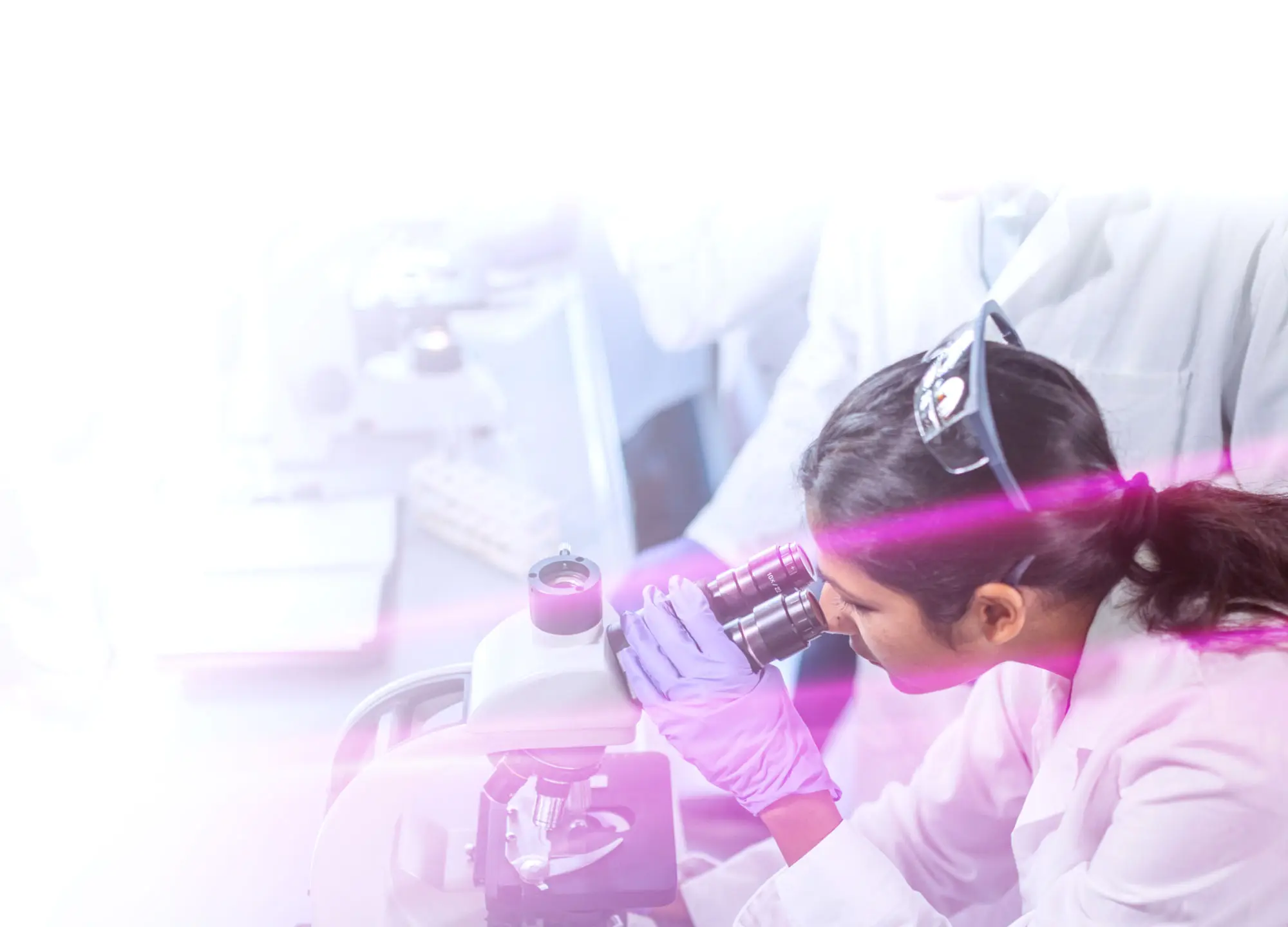 Scientist in a lab coat and gloves looking into a microscope; blurred laboratory background with colleagues.