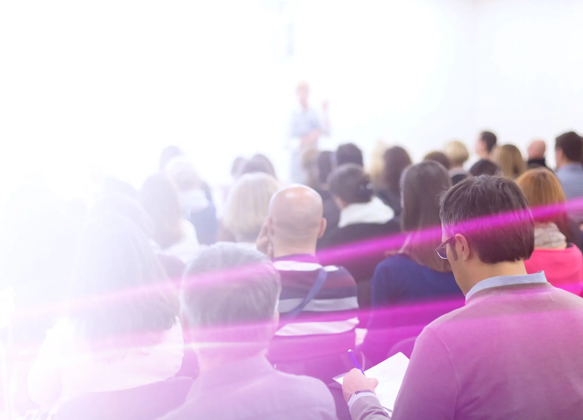 Blurred audience at a conference facing a speaker, with pink lens flare.